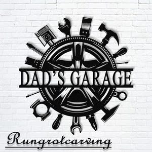 Puede incluir: Letrero de garaje de metal negro con el texto "DAD'S GARAGE". El letrero presenta un diseño circular con varias herramientas como llaves, martillos y destornilladores dispuestos alrededor de un diseño de rueda central. El letrero está montado en una pared de ladrillos blancos.