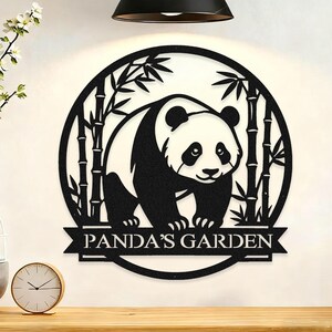 Op de afbeelding: Zwart en wit metalen wanddecoratie met een panda omringd door bamboe. De panda staat in een cirkelvormig frame, met de tekst "PANDA'S GARDEN" op een banner. Geschikt voor binnen en buiten.