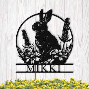 Könnte beinhalten: Schwarze Metall-Wandkunst mit einem Hasen in einem Kreis, umgeben von Blumen und Blättern. Der Name "MIKKI" ist darunter abgebildet. Das Kunstwerk ist vor einem weißen Holzhintergrund platziert.