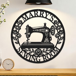 May include: Black metal wall art with a sewing machine design inside a circular frame. The words "MARRY'S SEWING ROOM" are cut out around the machine, with floral accents. A decorative piece for a sewing room, suitable for a craft space.