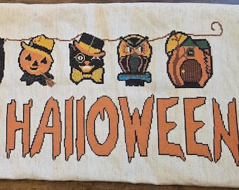 Peut inclure: Une bannière rectangulaire en tissu beige avec un motif au point de croix représentant une guirlande d'icônes d'Halloween, dont des citrouilles, une sorcière, un chat, un hibou et une maison. Le texte HAPPY HALLOWEEN est brodé en orange.