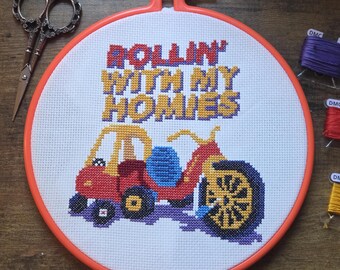 Peut inclure: Un projet de point de croix dans un tambour orange représentant une voiture jouet rouge et jaune avec le texte "ROLLIN' WITH MY HOMIES" en jaune et bleu. À côté du tambour se trouvent des ciseaux en métal et trois bobines de fil violet, rouge et jaune.
