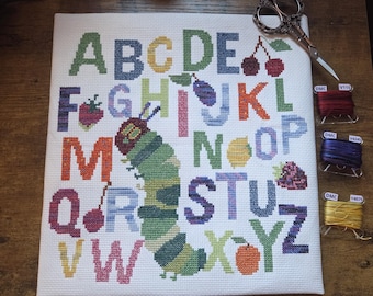 Peut inclure: Un échantillon de point de croix avec l'alphabet sur tissu blanc, illustré d'une chenille verte et de divers fruits. Le motif comprend des lettres colorées de A à Z, des ciseaux en métal de style ancien et trois bobines de fil DMC.
