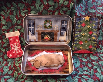 Peut inclure: Ensemble d'articles de Noël faits à la main en point de croix. Comprend une chaussette rouge et blanche, une scène miniature dans une boîte avec une cheminée et un motif de sapin de Noël. Les articles sont sur un tissu à motifs de houx.