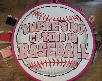 Peut inclure: Un projet de point de croix dans un tambour à broder rose représentant un motif de baseball avec des coutures rouges et le texte THERE'S NO CRYING IN BASEBALL en lettres roses et rouge foncé, avec trois petites bobines de fil à broder.