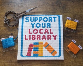 Peut inclure: Broderie au point de croix avec le texte "SUPPORT YOUR LOCAL LIBRARY" en bleu et rose. L'œuvre présente une bordure bleue et des images de livres. Inclut du fil à broder bleu, orange et jaune, et des ciseaux ornés.