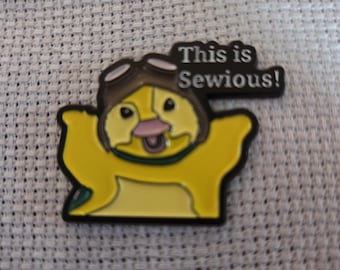 Peut inclure: Épingle en émail jaune représentant un canard de dessin animé portant un casque et des lunettes d'aviateur bruns. L'épingle porte le texte "This is Sewious!" en noir. L'épingle est bordée de noir.