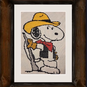 May include: Cross-stitch artwork of Snoopy in a cowboy hat, red bandana, and vest, holding a rifle. The artwork is framed in a rustic, dark wood frame, with a white matting. The Snoopy image is on a cream-colored background.