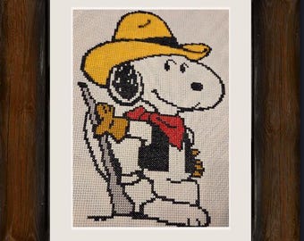 Peut inclure: Broderie au point de croix de Snoopy portant un chapeau de cowboy, un bandana rouge et un gilet, tenant un fusil. L'œuvre est encadrée dans un cadre en bois foncé rustique, avec un passe-partout blanc. L'image de Snoopy est sur fond crème.
