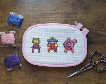 Peut inclure: Un projet de point de croix avec trois personnages pixélisés colorés dans un cercle ovale rose. Du fil à broder bleu, rose et violet est à côté du cercle, avec une paire de ciseaux décoratifs. Le projet est sur une surface en bois.