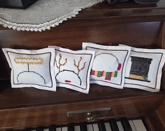 Peut inclure: Quatre ornements de Noël blancs brodés au point de croix avec des boucles de suspension rouges. Les motifs comprennent un ange, un renne, un bonhomme de neige et un haut-de-forme. Chaque ornement a une bordure marron.