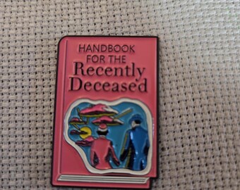 Peut inclure: Une épingle en émail rose en forme de livre avec le titre "Handbook for the Recently Deceased". L'épingle présente une illustration colorée de deux personnages et de soucoupes volantes. L'épingle a une bordure noire.