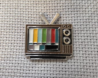 Peut inclure: Une épingle en émail argenté en forme de téléviseur rétro. L'écran affiche un motif de test coloré. L'épingle a deux antennes et deux boutons circulaires sur le côté. L'épingle est posée sur un fond texturé gris clair.