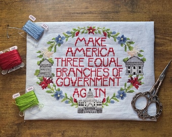 Peut inclure: Une broderie au point de croix avec le texte "MAKE AMERICA THREE EQUAL BRANCHES OF GOVERNMENT AGAIN" en rouge, entourée d'une couronne florale. Inclut des bobines de fil DMC bleu, rouge et vert, et une paire de ciseaux.