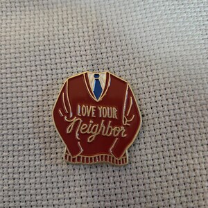 May include: A red enamel pin in the shape of a sweater with a blue tie. The pin has the words "LOVE YOUR Neighbor" in a cursive font. The pin is on a light gray textured surface.