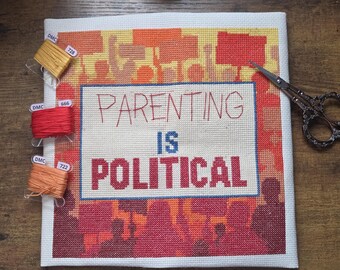 Peut inclure: Un projet de point de croix sur tissu blanc avec le texte "PARENTING IS POLITICAL" en rouge et bleu, entouré d'un fond de silhouettes orange et rouges de personnes tenant des pancartes. Trois bobines de fil jaune, rouge et orange et des ciseaux.