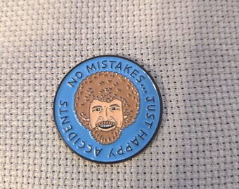 Peut inclure: Épingle ronde en émail bleu représentant un visage souriant avec une afro et une barbe brunes. L'épingle porte l'inscription "NO MISTAKES... JUST HAPPY ACCIDENTS" en lettres blanches sur le bord. L'épingle est posée sur un fond texturé bleu clair.