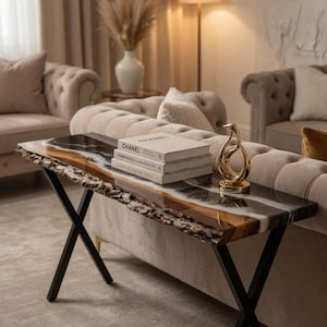 May include: A modern live edge console table with a black metal base. The tabletop features a unique design with brown, white, and black resin. Three books and a gold sculpture are displayed on the table.