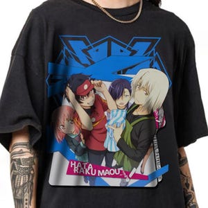 May include: Black t-shirt featuring a colorful anime graphic. The graphic includes multiple characters with varying hair colors, set against a blue background. The text "HATA RAKU MAOU" is visible at the bottom of the graphic.