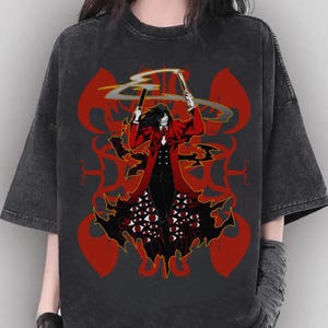 May include: Dark gray t-shirt featuring a graphic of a character in a red coat holding two weapons. The character is surrounded by red, stylized shapes and a black base with multiple eyes. The shirt has short sleeves.