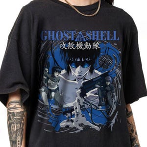 May include: Dark gray t-shirt featuring a graphic from the anime series "Ghost in the Shell." The design includes the title text in blue and Japanese characters, along with multiple anime characters in action poses, set against a blue and gray background.