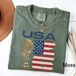 May include: Moss green t-shirt with a distressed "USA 2026" graphic. The graphic features the text "USA" in blue, a trophy, and a stylized American flag design. A gold necklace rests on the shirt.