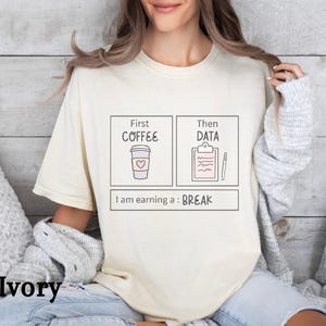 May include: An ivory short-sleeve t-shirt featuring a graphic with two boxes labelled First COFFEE with a cup icon and Then DATA with a clipboard and pen icon. Below, the text reads I am earning a : BREAK. The shirt is made of cotton fabric.