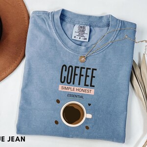 May include: A blue jean colored t-shirt featuring the text COFFEE, SIMPLE HONEST, and ESSENTIAL printed in black and white. Below the text is a graphic of a coffee cup surrounded by small brown coffee beans. The shirt has a Comfort Colors label.