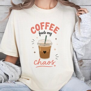 May include: A cream-coloured short-sleeve t-shirt featuring a graphic of an iced coffee in a plastic cup with a straw. The text "COFFEE fuels my chaos" is printed in coral and black lettering above and below the coffee cup illustration.