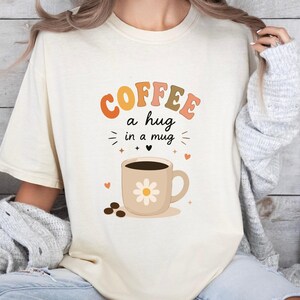 May include: A cream-colored t-shirt featuring a graphic of a coffee mug with a white daisy design. The text above the mug reads "COFFEE a hug in a mug" in orange, pink, and brown letters, accompanied by small heart and star icons and coffee beans.