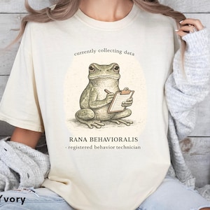 May include: An ivory short-sleeve t-shirt featuring a graphic of a frog holding a clipboard and pen. The text reads currently collecting data, RANA BEHAVIORALIS, and registered behavior technician. The frog is illustrated in shades of green and brown.