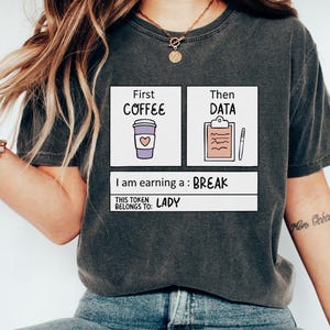 May include: Dark grey t-shirt featuring a graphic with the text "First Coffee, Then Data, I am earning a Break." The design includes a coffee cup with a heart and a clipboard with a pen. The text "This token belongs to: Lady" is also included.