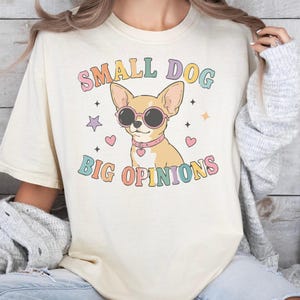 May include: A cream-colored t-shirt featuring a graphic of a tan chihuahua wearing pink-rimmed sunglasses and a pink collar with a heart charm. The text "SMALL DOG BIG OPINIONS" is printed in colorful, retro-style lettering, surrounded by small stars and hearts.