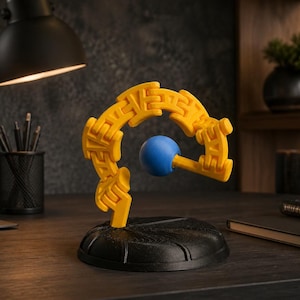 May include: A yellow articulated plastic chain structure suspended in a curved arch shape, featuring a solid blue sphere attached to the center. The entire assembly is mounted on a circular black base with a textured surface and radial grooves.