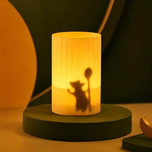 May include: A cylindrical table lamp with a warm, glowing light. The translucent shade features a dark silhouette of a small creature holding a long, round-topped staff. The lamp has a vertical ribbed texture and emits a soft, diffused yellow light.