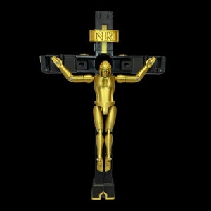 May include: A gold-coloured articulated figure on a black cross with the letters INRI in gold. The figure has jointed arms and legs, and the cross has a modern, geometric design. The background is solid black.