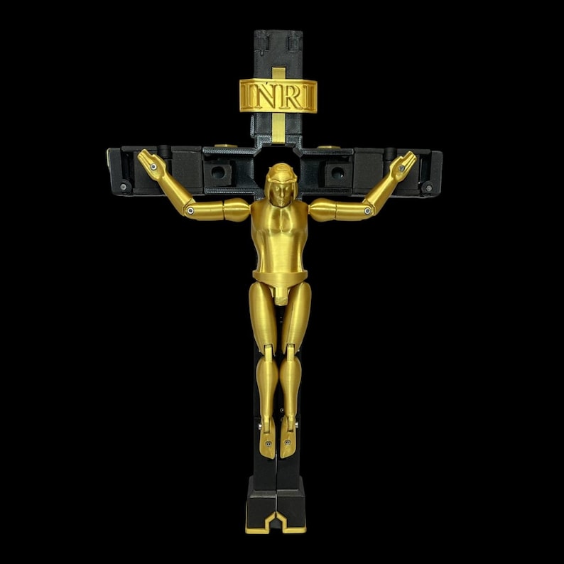 May include: A gold-coloured articulated figure on a black cross with the letters INRI in gold. The figure has jointed arms and legs, and the cross has a modern, geometric design. The background is solid black.