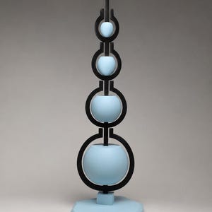 Può includere: Una scultura verticale con quattro sfere azzurre di dimensioni decrescenti, ciascuna inserita in una cornice circolare nera. La struttura è sostenuta da una base azzurra e da un piccolo blocco quadrato, creando una disposizione impilata.