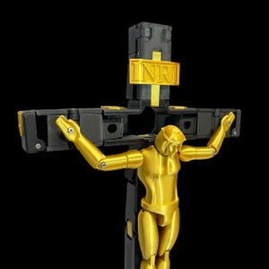 May include: A gold-coloured articulated figure on a black cross with a gold INRI sign. The figure has its arms outstretched. The cross is black with gold accents. The background is black.
