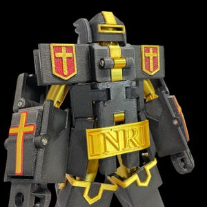 May include: A black and gold robot figure with religious symbols. The robot has a helmet, shield, and chest plate with a cross design. The chest plate has the letters "INRI" in gold. The robot's joints and accents are gold.