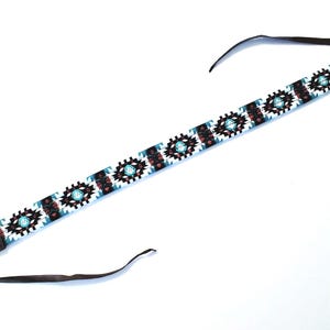 May include: A long, narrow camera strap featuring a repeating geometric pattern in white, black, turquoise, and dark brown. The central section is decorated with beadwork, while the ends are finished with dark brown leather tabs and thin straps.