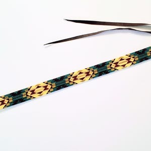 May include: A long, narrow strap featuring a repeating geometric pattern in shades of yellow, dark green, and maroon. The strap is finished with dark brown leather tabs at both ends, with one end featuring thin, dark brown leather ties for attachment.
