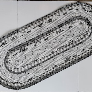 May include: Oval braided rug in shades of gray and white. The rug features a mix of patterns, including gingham and other designs. The rug is made of fabric strips and is suitable for a hallway or entryway.