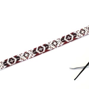 May include: A long, narrow guitar strap featuring a repeating geometric pattern in shades of deep maroon, black, and white. The strap ends are finished with dark brown leather tabs, each containing a metal grommet and thin black leather ties.