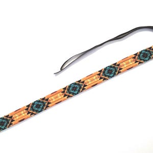 May include: A long, narrow camera strap featuring a repeating geometric pattern in shades of orange, black, and turquoise. The strap is finished with dark brown leather end tabs, each secured with a metal rivet, and includes a thin black cord.