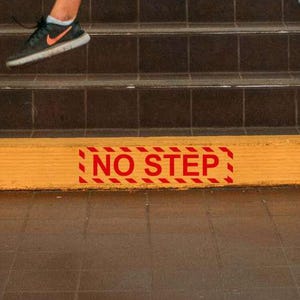 May include: A person jumping on a staircase, holding a camera. The stairs are brown with yellow accents and a "NO STEP" warning. The person is wearing a black shirt, olive green trousers, and black trainers.