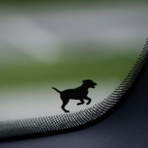May include: Black silhouette of a dog in mid-stride, appearing to run. The dog is positioned on a curved surface with a mesh pattern, set against a blurred green and gray background. The image suggests a decal or sticker.
