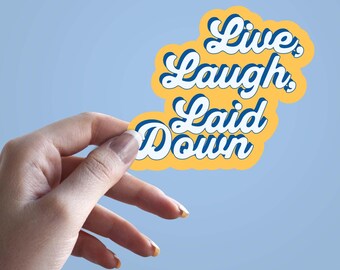 Pode incluir: Um autocolante amarelo com as palavras "Live, Laugh, Laid Down" numa fonte retro com detalhes azuis e brancos. O autocolante é segurado por uma mão sobre um fundo azul claro.