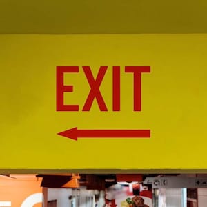 May include: A yellow sign with the word "EXIT" in bold red letters and a red arrow pointing left. The sign is mounted on a yellow surface, indicating the direction of an exit.