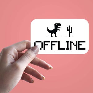 May include: A white rectangular sticker with a black pixelated dinosaur and cactus graphic, with the word "OFFLINE" in black pixelated letters. The sticker is held against a pink background.